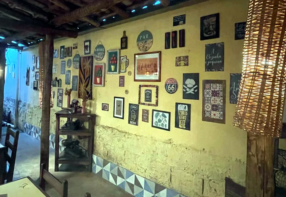 A parede amarela do salão, cheia de quadros, com as placas redondas de Paz, Saúde e Amor e o barrado de azulejo
