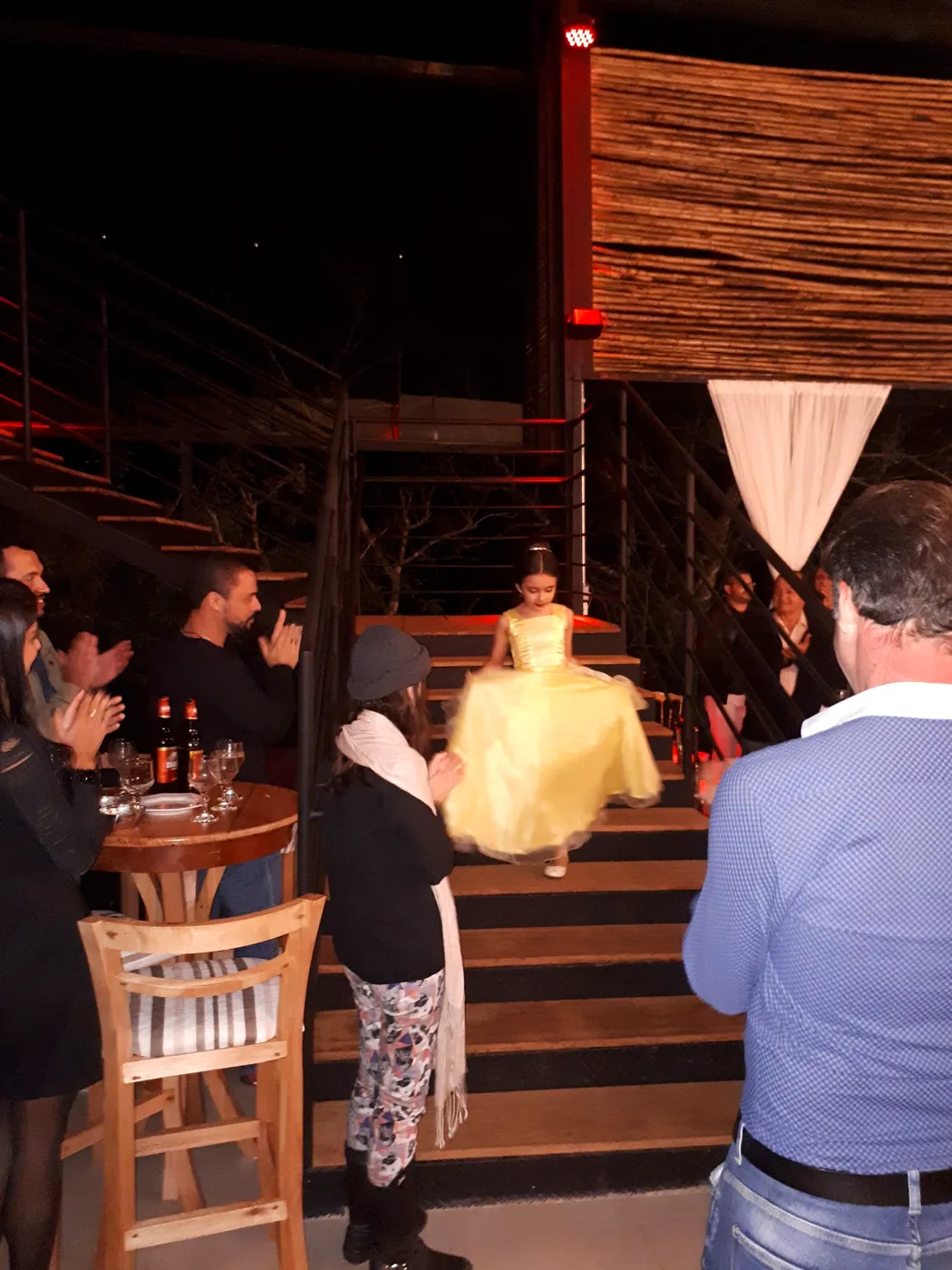 Debutante de vestido amarelo no alto da escada do salão, com convidados aplaudindo