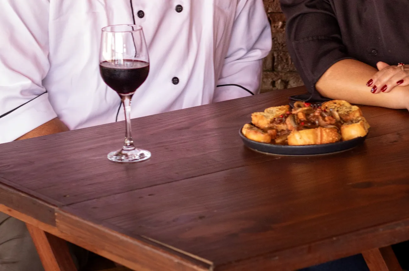Mesa da Em Cantos com taça de vinho, a garrafa e uma porção, na frente da parede de tijolinho