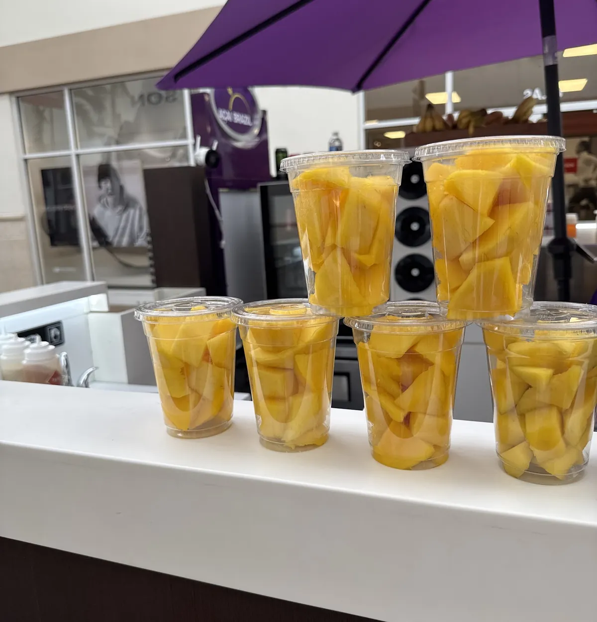 Cups of fresh mango lined up on the Flow Açaí kiosk counter, under the purple umbrella